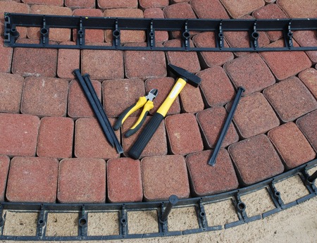 Zdjęcie przedstawia leżace na kostce ogrodowe obrzeże plastikowe, kotwy do mocowania obrzeża, młotek i szczypce. Na dole obrazka widać ułożone na podbudowie, wygięte w łuk obrzeże plastikowe z wbitą kotwą